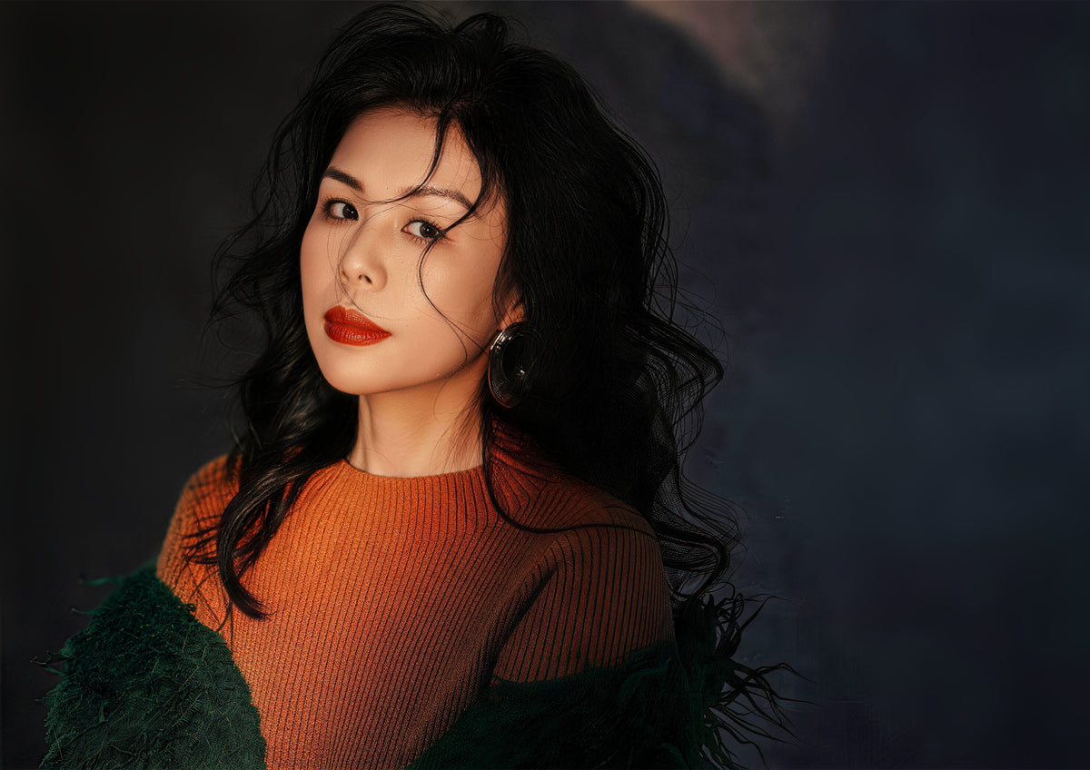 A woman with dark wavy hair wearing an orange top against a dark background.
